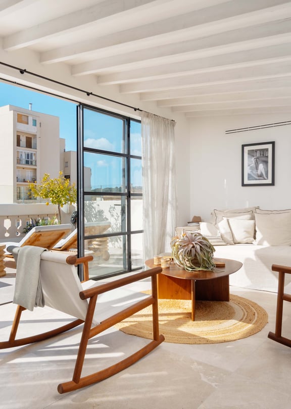Upper-level sitting room with wooden rocking chairs and a cream sofa, steel-framed doors open to the terrace and rooftops beyond.