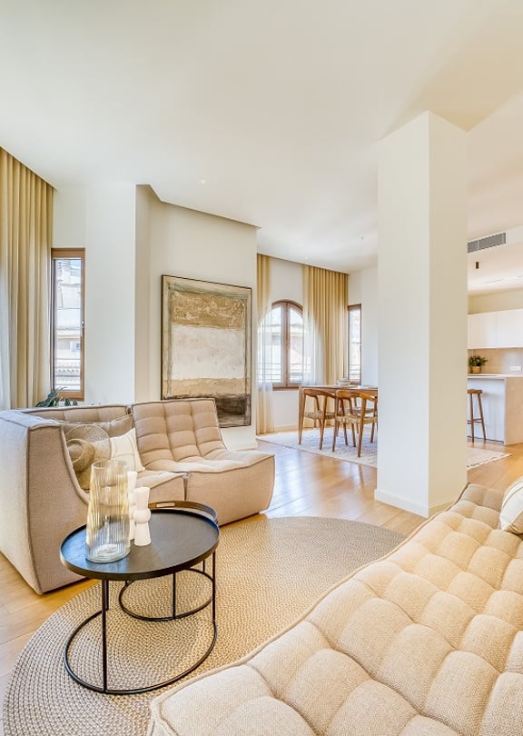 A bright living room in a penthouse with arched windows, light oak flooring, beige modular sofas, and a dining area in the background.