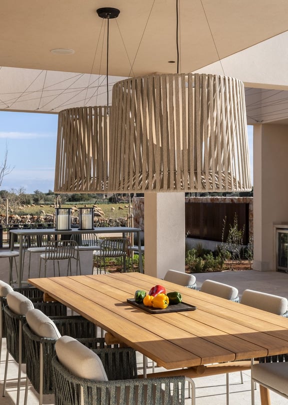 A spacious outdoor dining patio features a long wooden table, woven chairs, and a stone-built summer kitchen, looking towards mountain fields.