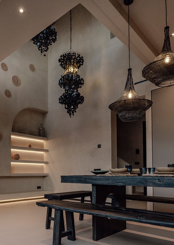 Dining area featuring a dark wooden farmhouse table and benches, sculptural black pendant lights, and arched wall niches with soft integrated illumination.