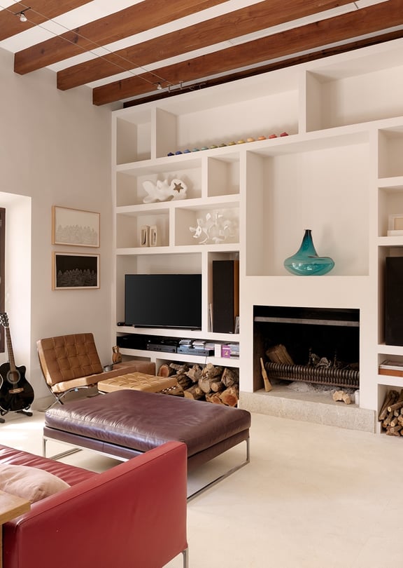 Living room with exposed wood ceiling beams, white built-in shelving unit, dark red sofa, and an open fireplace.
