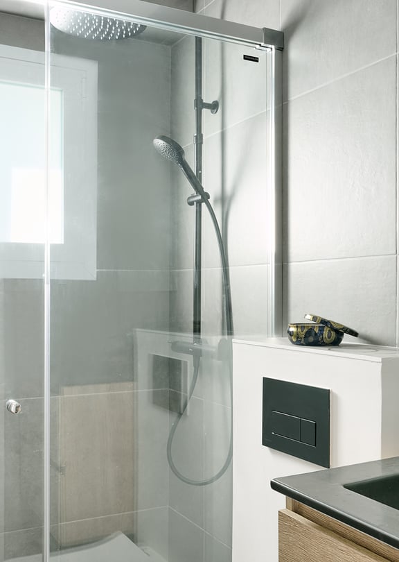 A bathroom with grey tiled walls, a glass-enclosed shower with a rainfall head, and a vanity with a black sink.