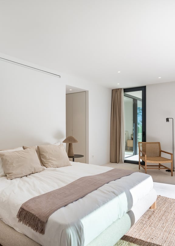 Serene master bedroom with neutral linen bedding, rattan accent chair and floor-to-ceiling sliding doors to a private terrace with pine tree views