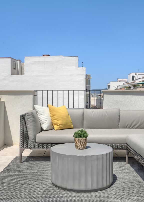 A rooftop terrace in El Molinar featuring a grey modular sofa with yellow cushions, a round fluted concrete table, and a patterned rug against a bright sky.