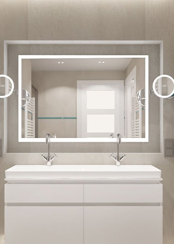 A modern bathroom featuring a white double vanity, large rectangular mirror with integrated lighting, and white heated towel rails against neutral stone-tiled walls.
