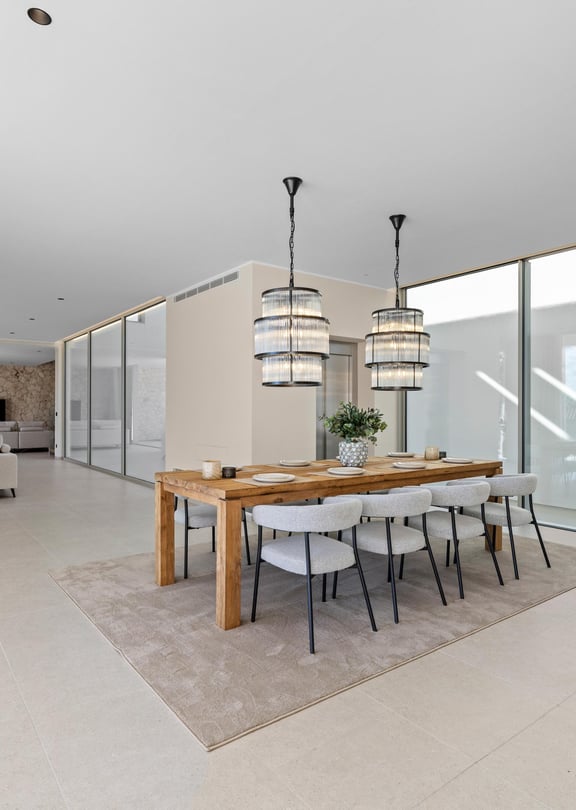 Open-plan living and dining area featuring floor-to-ceiling windows, a wooden dining table, fabric chairs, and designer lighting.