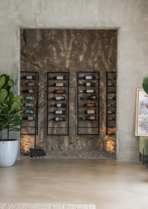 Minimalist wine storage area featuring black metal racks against an exposed rock wall and raw concrete finishes.