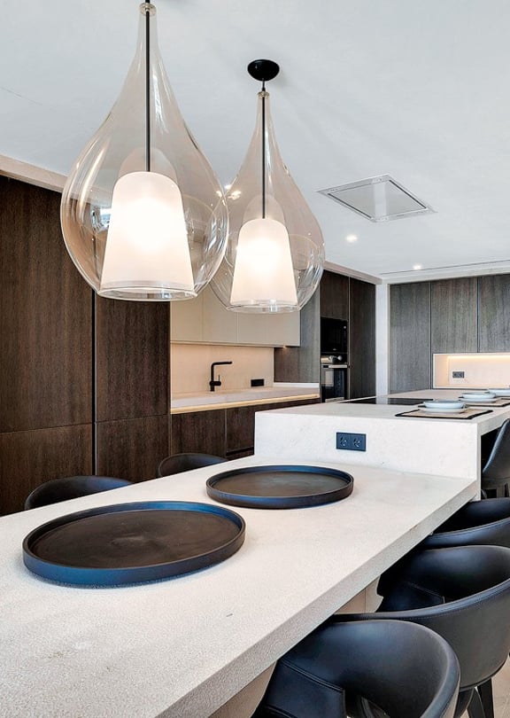 Modern open-plan kitchen featuring a long light-colored stone island, dark cabinetry, and two clear glass pendant lights.