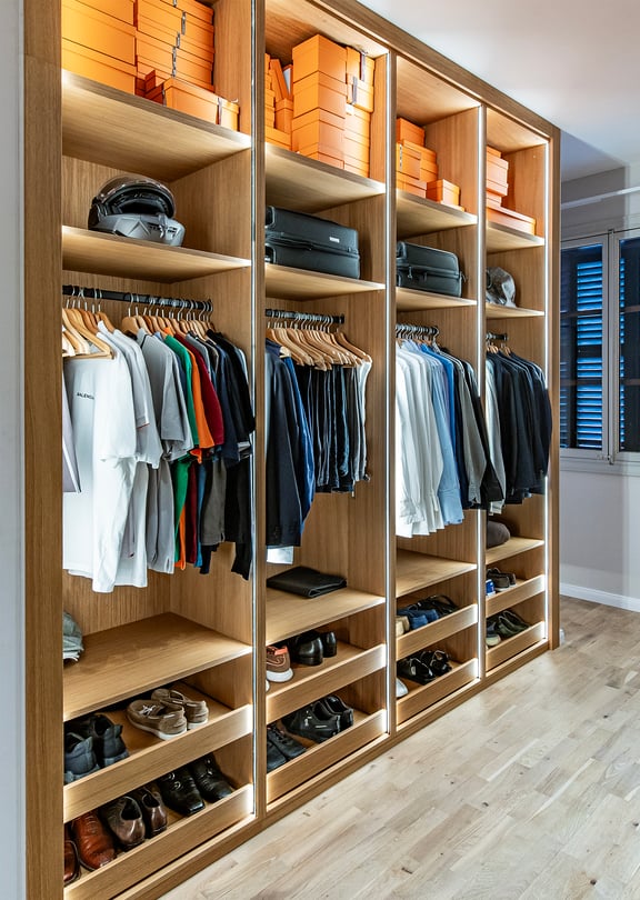 A walk-in dressing room featuring light oak cabinetry with illuminated shelving, hanging rails, and shoe racks, leading to an adjacent bedroom.