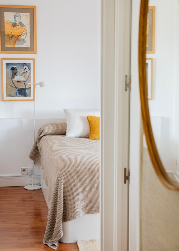 A close-up view of a bedroom interior with a bed featuring textured bedding, two framed artworks on the white wall, and a large oval mirror in the foreground.