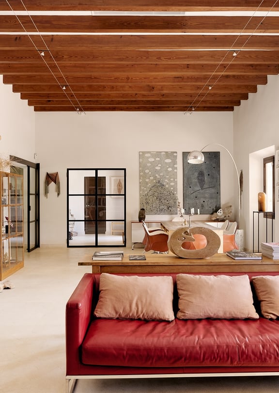 A spacious living room with high beamed ceilings, red leather sofa, and white walls in a Finca-style home.