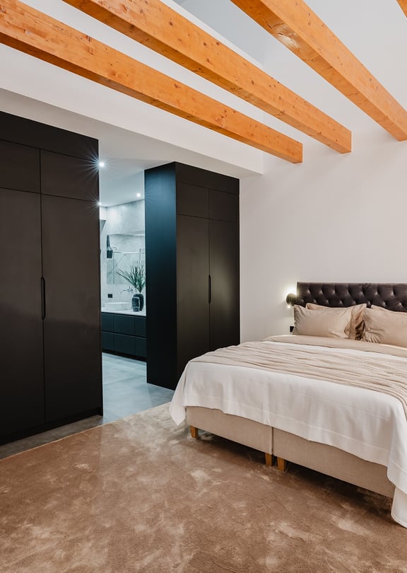 A modern bedroom with a large bed against a white wall, dark floor-to-ceiling wardrobes, and visible natural wood ceiling beams.