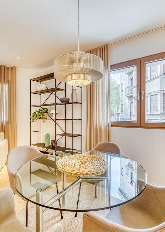 A glass dining table with four curved chairs next to a metal shelving unit in an apartment, with linen curtains framing city windows.