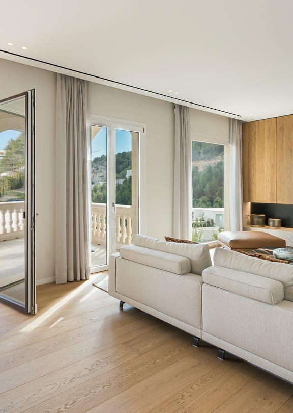 Spacious living room with beige sofa, wooden flooring, and floor-to-ceiling glass doors opening to a stone-railed terrace.