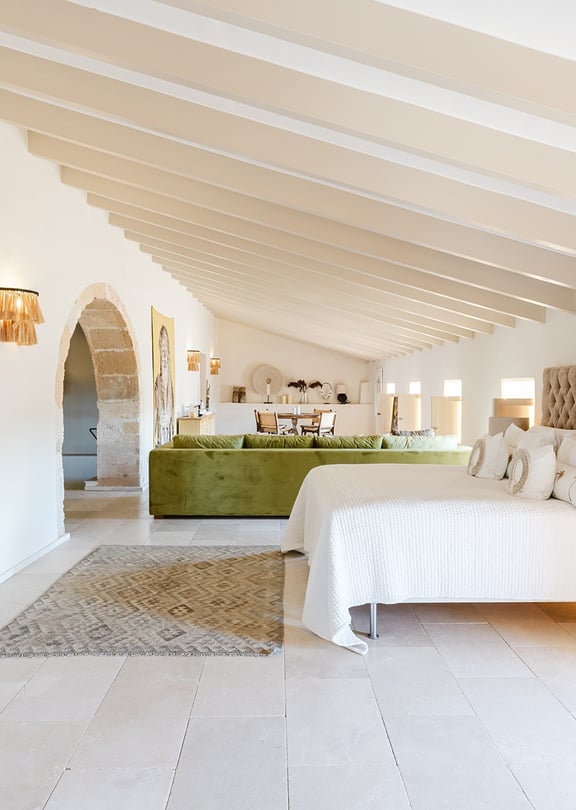 A spacious bedroom with a king-sized bed, tufted headboard, stone flooring, and arched doorways leading to an adjoining area.