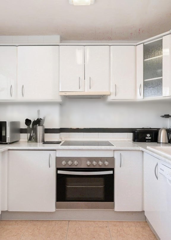 A bright galley kitchen with white cabinets, light countertops, and dark tiled backsplash, illuminated by natural light.