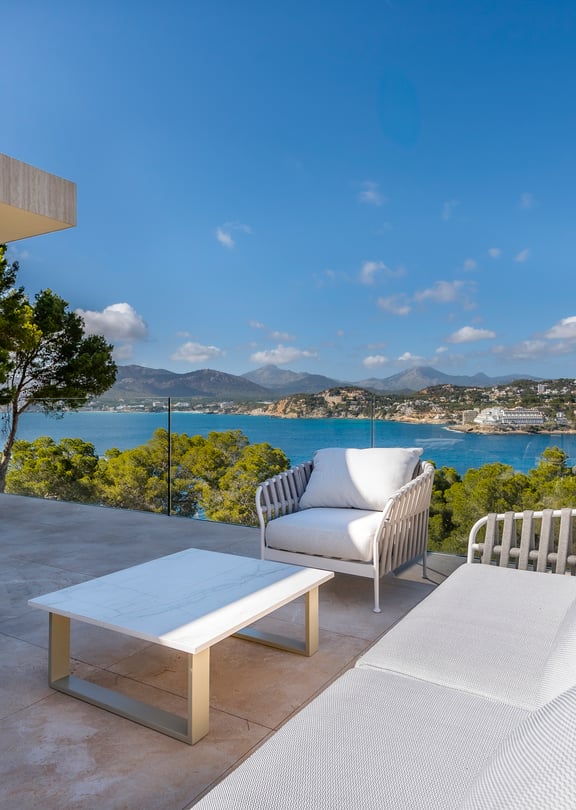 Outdoor terrace with white lounge furniture, glass railings, and a wide view of the Mediterranean bay and coastal mountains.
