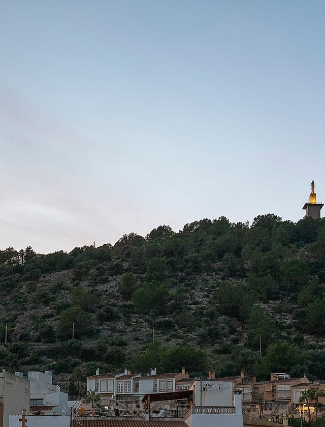 A distant view of a hillside topped with a tall landmark tower against a clear blue evening sky, seen above local rooftops.