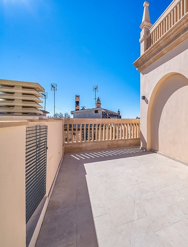 A rooftop terrace featuring light-colored stone flooring, stucco walls with a decorative arch, and stone balustrade railings under a clear blue sky.