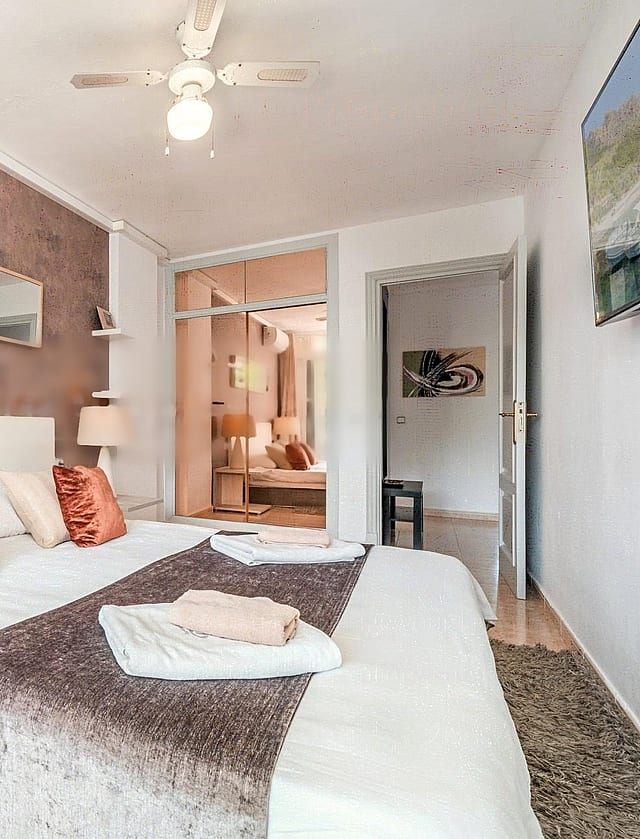 A bedroom featuring a textured dusty rose accent wall, a white bed with orange decorative pillows, a ceiling fan, and a view into a secondary room through an open doorway.