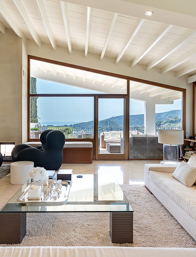 Spacious villa living room with white painted beams, fireplace, marble floors, designer seating and glazed wall with sea views