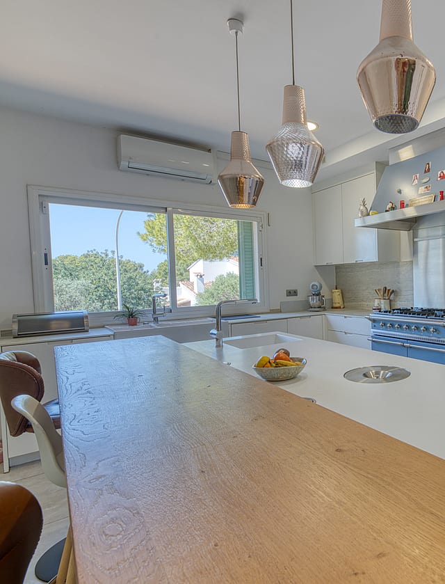 A modern white kitchen featuring a large central island with a wooden breakfast bar, stainless steel stove, and three glass pendant lights hanging above.