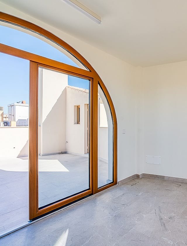 A minimalist room with white walls, stone flooring, and a large wooden arched glass door leading to an outdoor terrace.