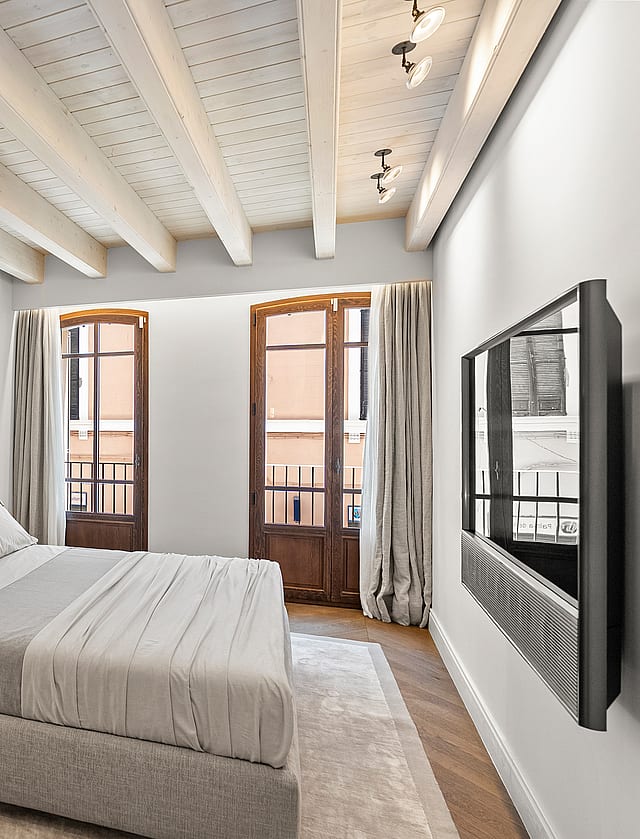 A bedroom featuring exposed timber ceiling beams, herringbone oak floors, and two arched glass doors opening to balconies.