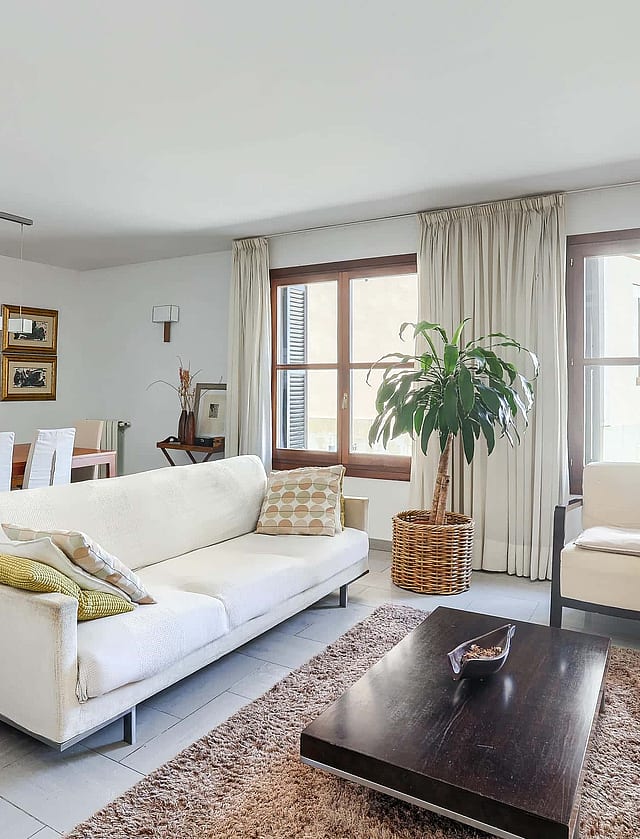A bright living room with a white sofa, dark wooden coffee table, and a dining table for six, featuring neutral decor.