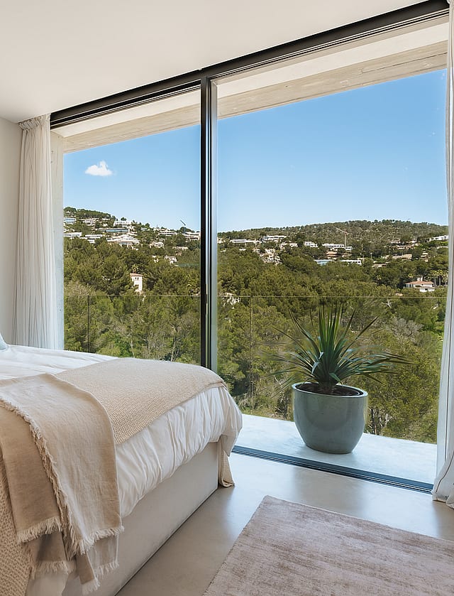 Modern bedroom with floor-to-ceiling windows overlooking green hills, featuring neutral textiles and a wooden side table.