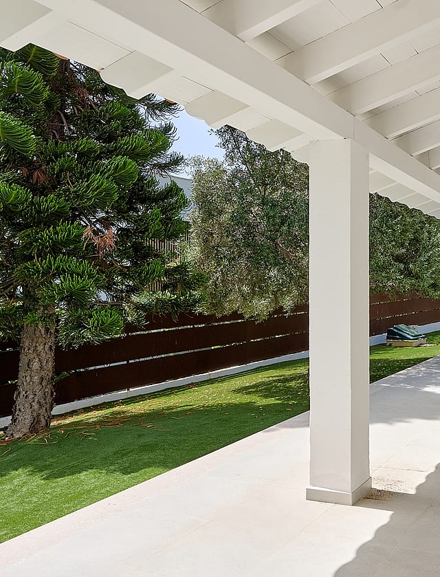 Terraza cubierta blanca con vigas de madera frente a césped artificial, piscina circular, pinos y cipreses