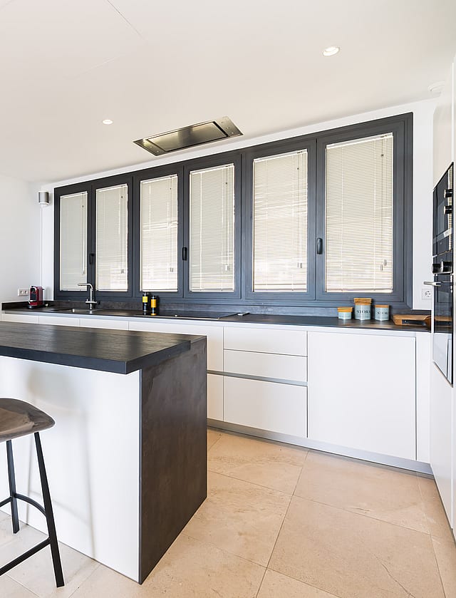 A minimalist kitchen featuring white handleless cabinetry, a dark grey island, and large dark-framed windows.