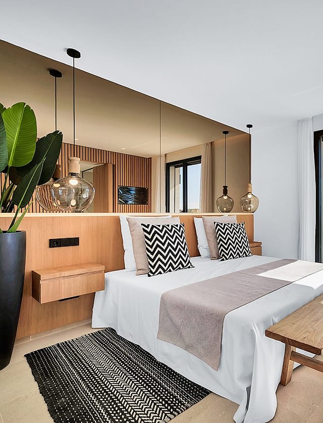 A modern bedroom featuring a bed with a wooden headboard, a large mirror, a wooden bench, and a floor-to-ceiling glass door.