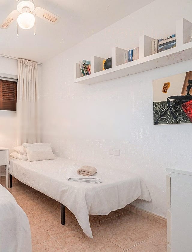 A bright guest bedroom featuring two single beds, light-toned textiles, a white wall-mounted shelf, and a minimalist aesthetic.