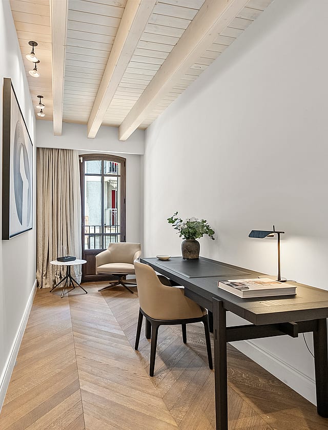 Minimalist home office featuring chevron wood flooring, exposed timber beams, a dark desk, and an arched glass door.