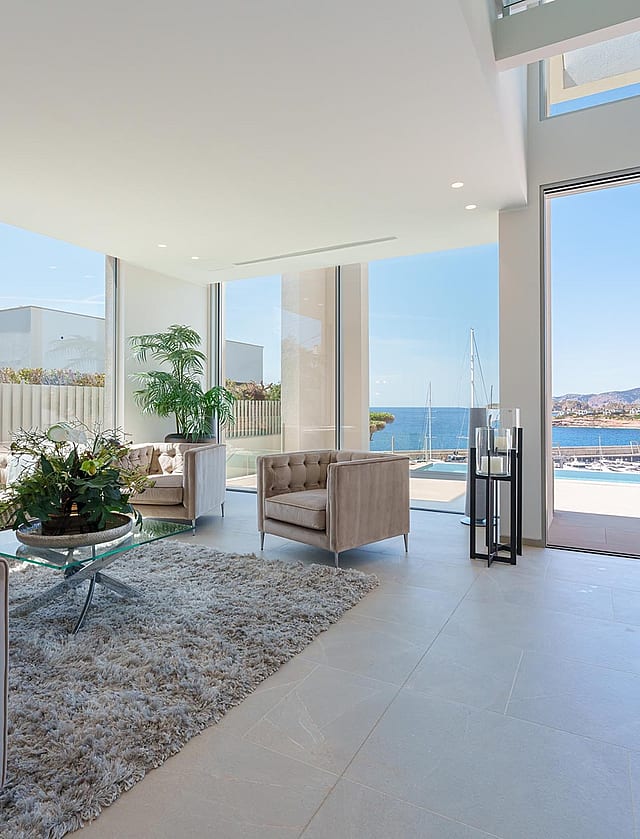 Bright living room with white walls, tufted beige sofas, glass coffee table, and large windows looking out to the harbor.