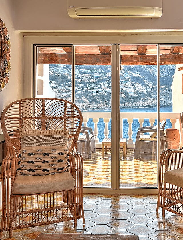 Two rattan armchairs facing sliding glass doors that open onto a sea-view terrace, with decorative mirrors and wall clock.