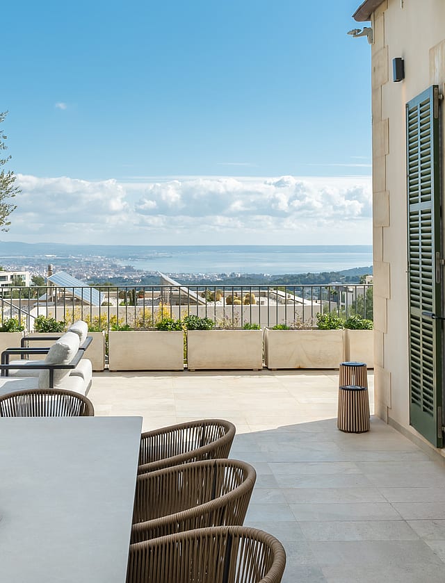 Outdoor dining terrace featuring a stone table, woven chairs, and a large olive tree overlooking the Son Vida landscape.
