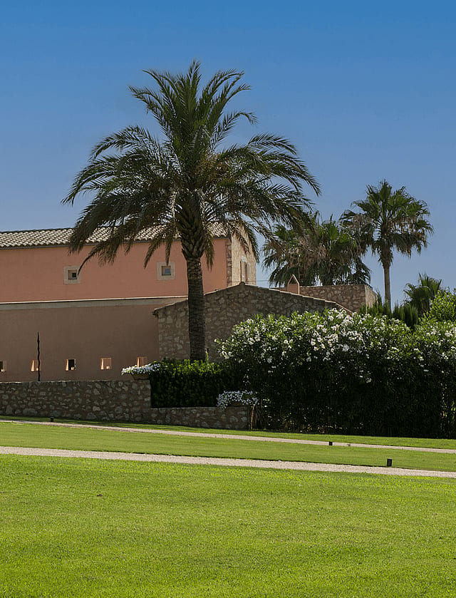 Exterior view of a traditional stone and peach-colored finca with arched entrance, large palm tree, and manicured green lawn.