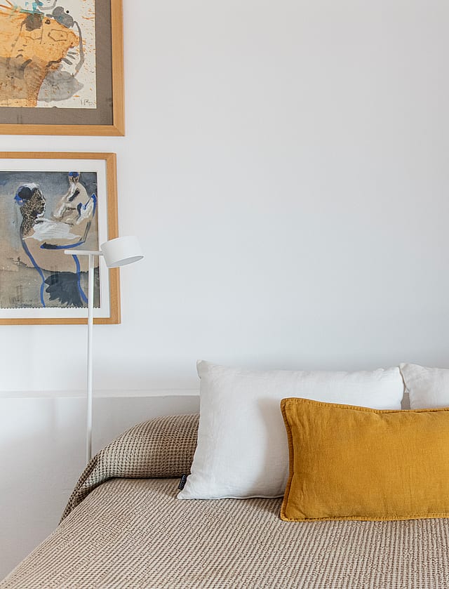 A double bed with a patterned beige cover and mustard yellow cushion, flanked by two minimalist floor lamps and framed wall art.