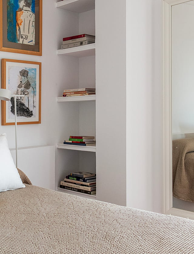 A close-up of a bedroom featuring a textured beige bedspread, a bright yellow throw pillow, and integrated white wall shelving.