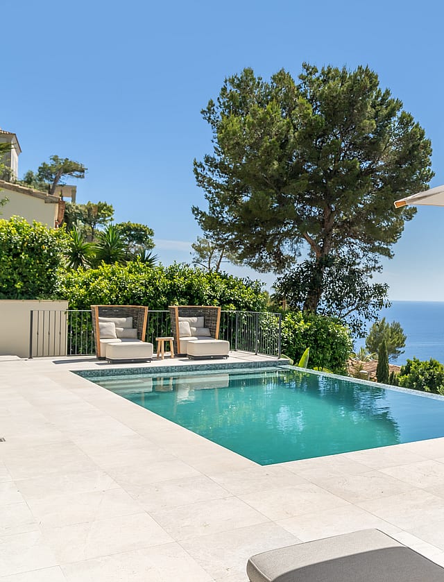 A rectangular swimming pool on a pale stone terrace overlooks the blue sea, framed by a pine tree and lounge furniture.