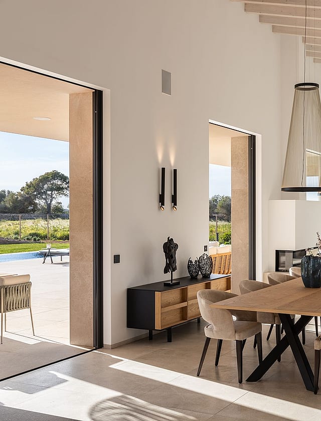 Dining room with a long oak table and cushioned chairs, open to an outdoor terrace, with white walls and exposed ceiling beams.