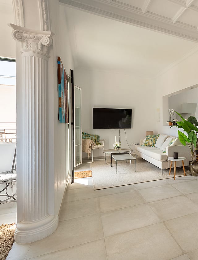 An open-plan living room in a Mediterranean apartment featuring a white column, stone tiled floors, a white sofa, and a kitchen visible in the background.