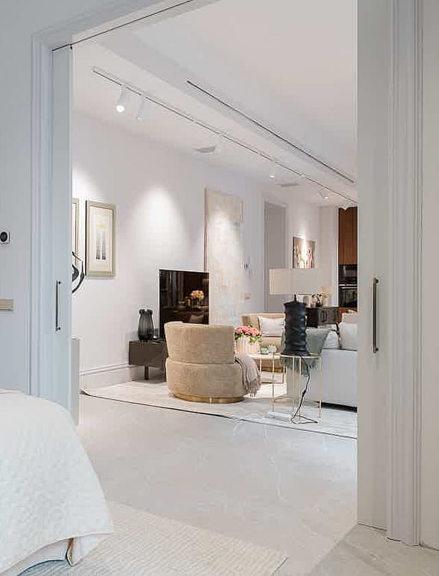 Interior view from a bedroom into an open-plan living room, featuring a beige headboard and neutral white decor.