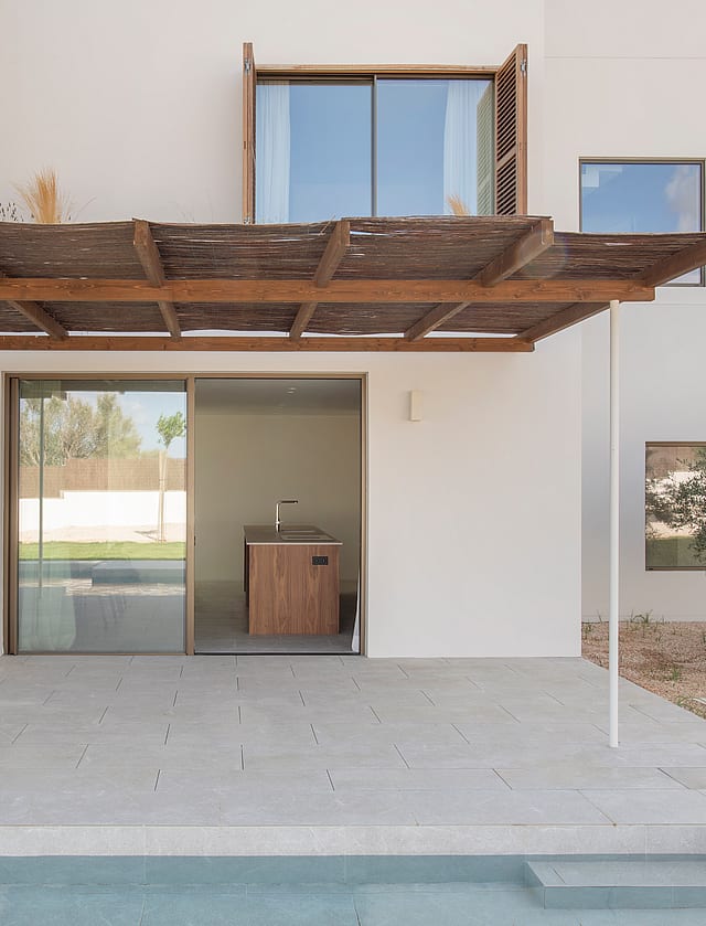 A stone terrace with a wooden pergola sits beside a swimming pool, leading to a modern white villa with glass doors.