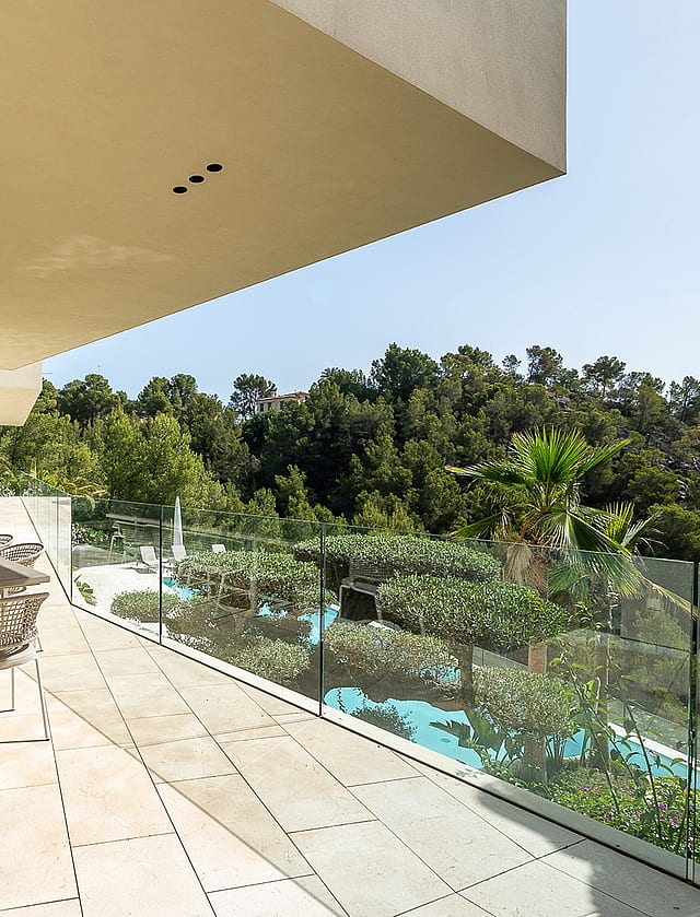 An expansive covered outdoor terrace featuring a dining table, natural stone flooring, and a glass balustrade overlooking trees.