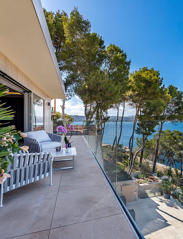 A wide terrace with grey outdoor furniture, glass railings, and a view overlooking the Mediterranean coastline and pines.