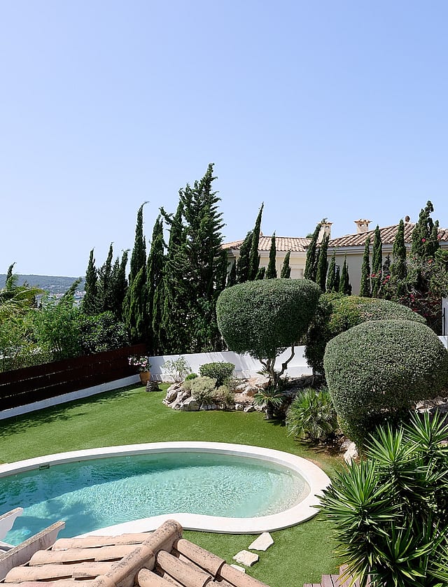 Vista desde tarima de madera sobre piscina ovalada, césped artificial, topiaria y palmeras con montañas al fondo