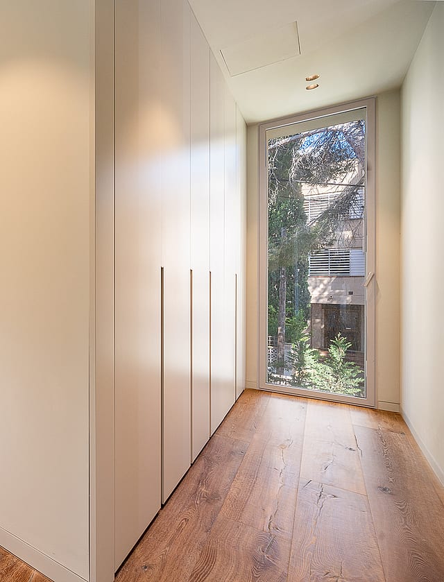 A minimalist dressing room corridor in a Santa Ponsa villa, featuring light oak wood floors, floor-to-ceiling white built-in wardrobes, and expansive glass windows overlooking greenery.